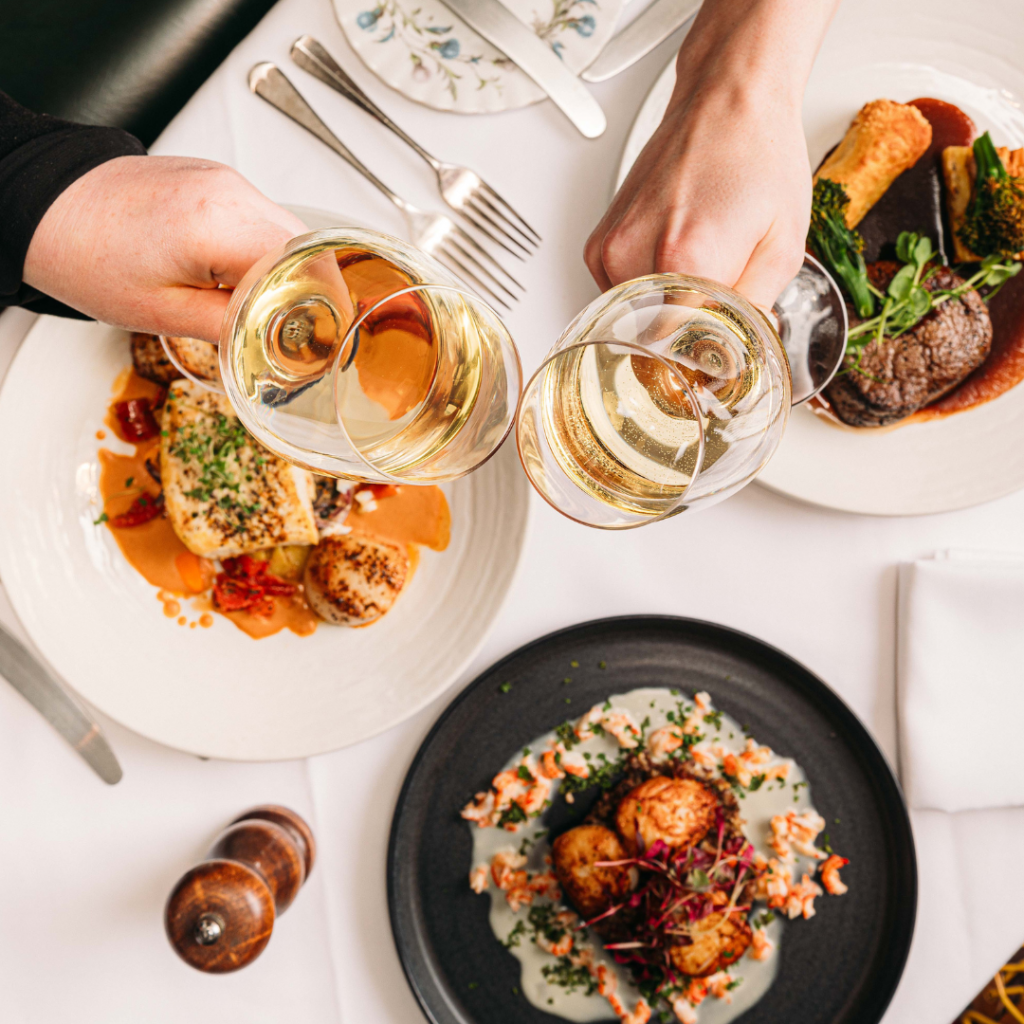 a table of food with two people clinking wine glasses over the top in a 'cheers' motion.