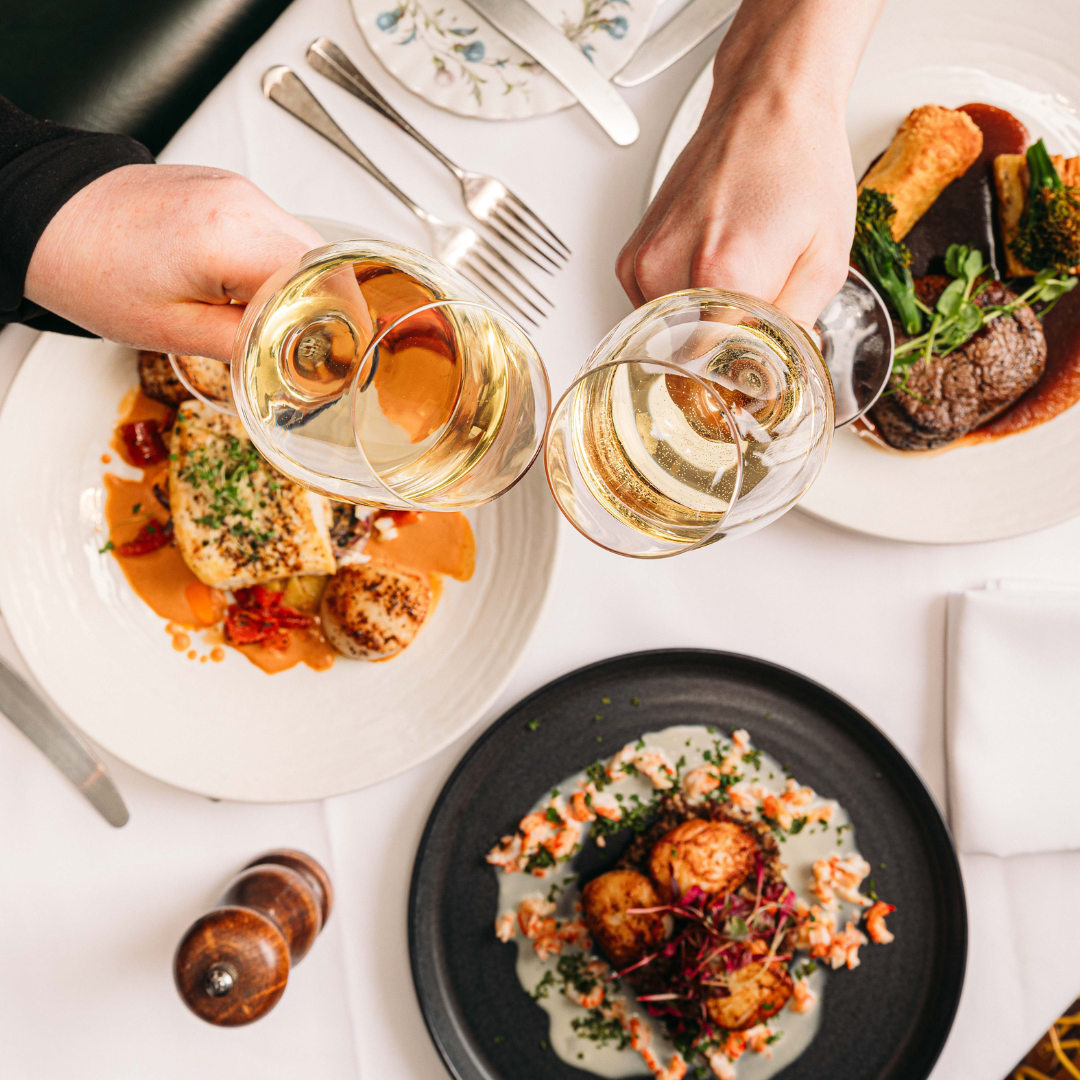 a table of food with two people clinking wine glasses over the top in a 'cheers' motion.